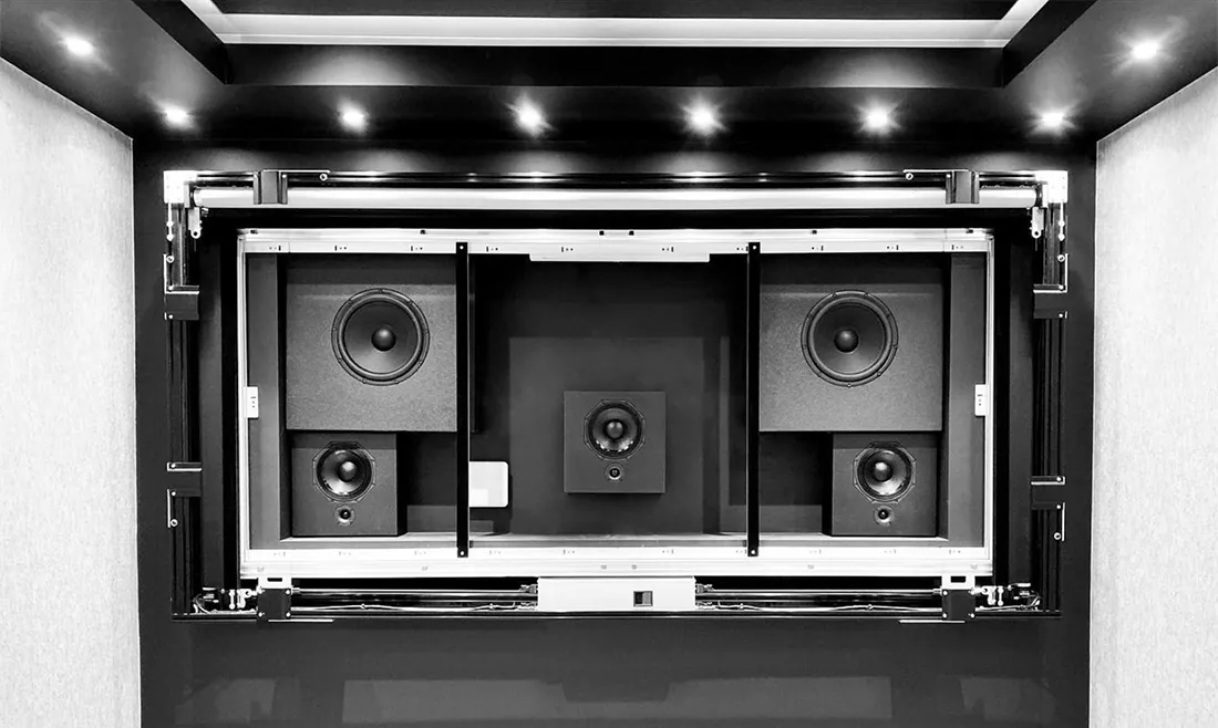 A cinema screen wall seen straight on: speakers set into a black frame where the screen will hang, lit by ceiling downlights.