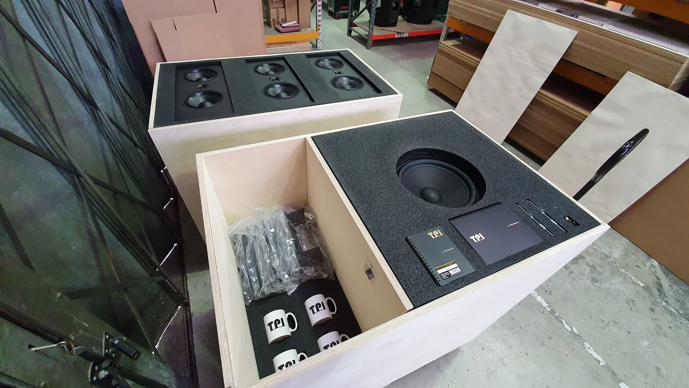 Open plywood crate with black foam inserts holding a loudspeaker and its accessories.