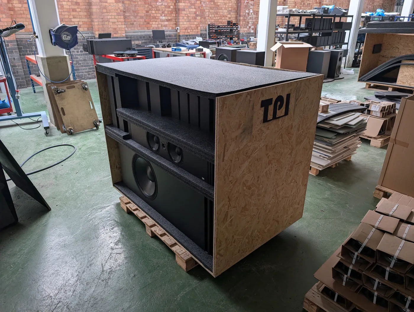 A plywood crate on a pallet in TPI's workshop with its front panel off, showing a screen speaker and a large subwoofer held between layers of black foam.