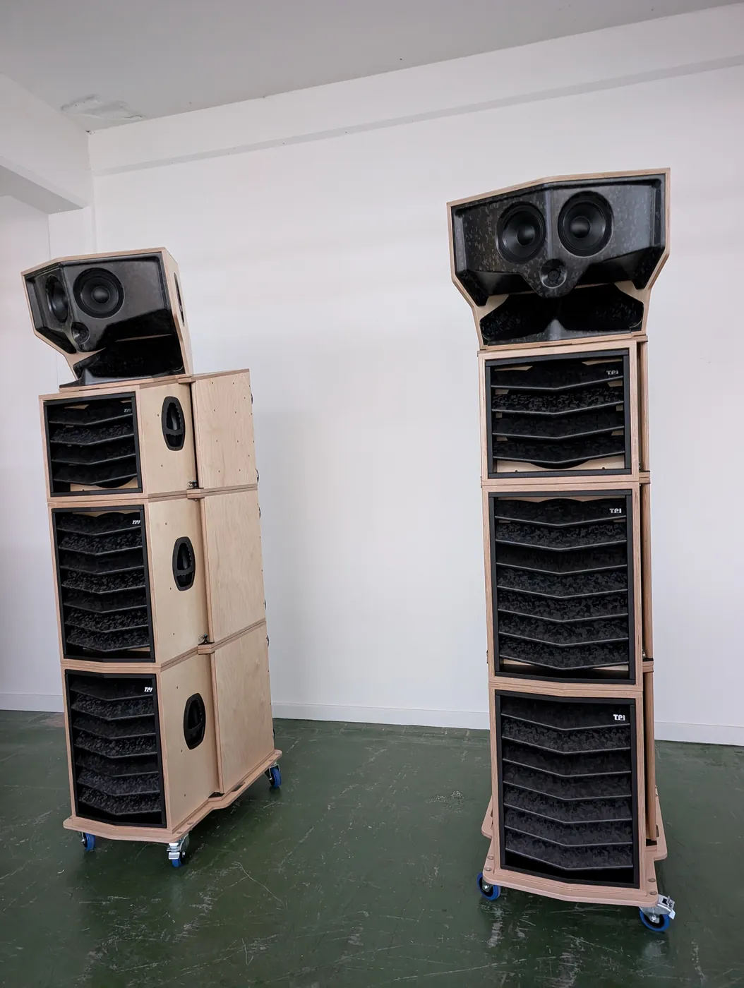 Two tall loudspeaker stacks in natural birch plywood on castor boards: slatted bass modules below angled horn-and-mid top sections, in a white room.