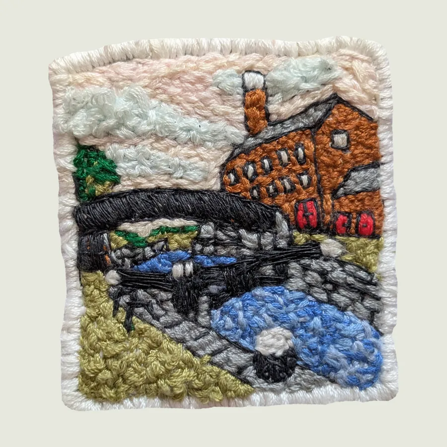 Square embroidered patch edged in white: an orange-brick building with a chimney beside a dark arched bridge over blue water, with green and grey banks.