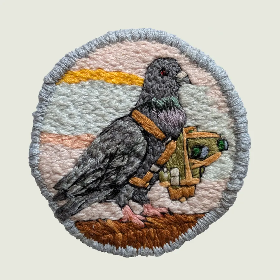 Round embroidered patch edged in pale grey: a grey pigeon beside a small yellow and green object, on an orange ground under a pale sky.