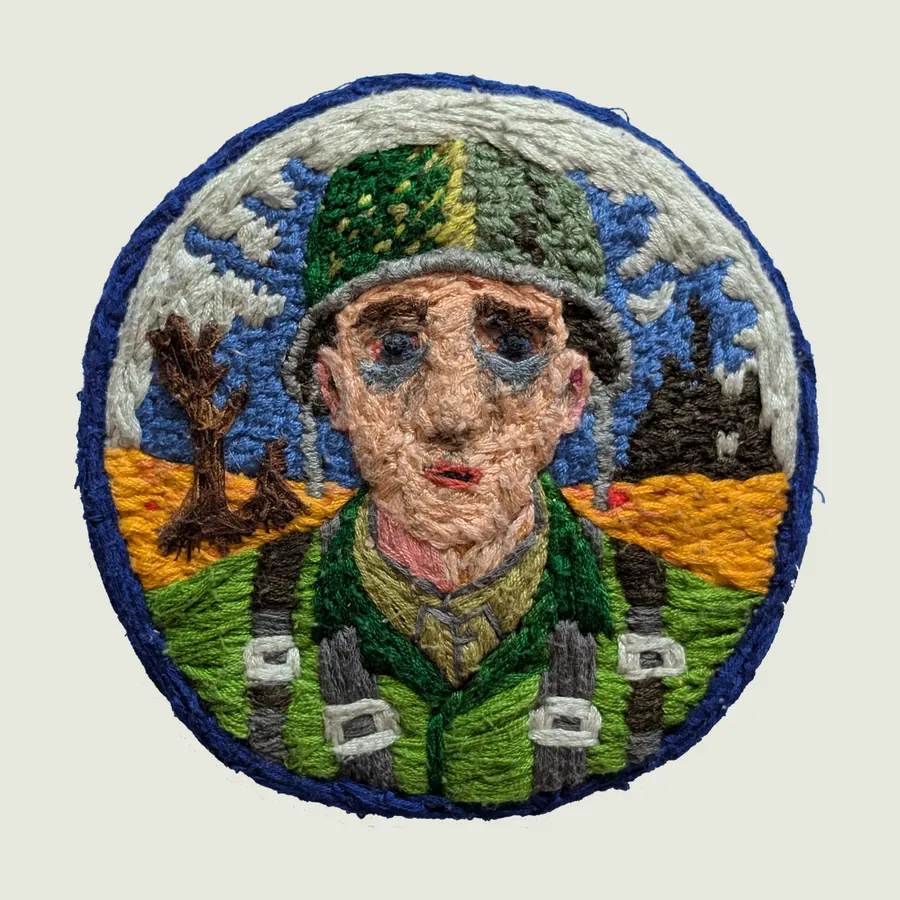 Round embroidered patch edged in blue: a soldier in a green helmet and green uniform with grey straps, in front of an orange landscape, a bare brown tree and a blue and white sky.
