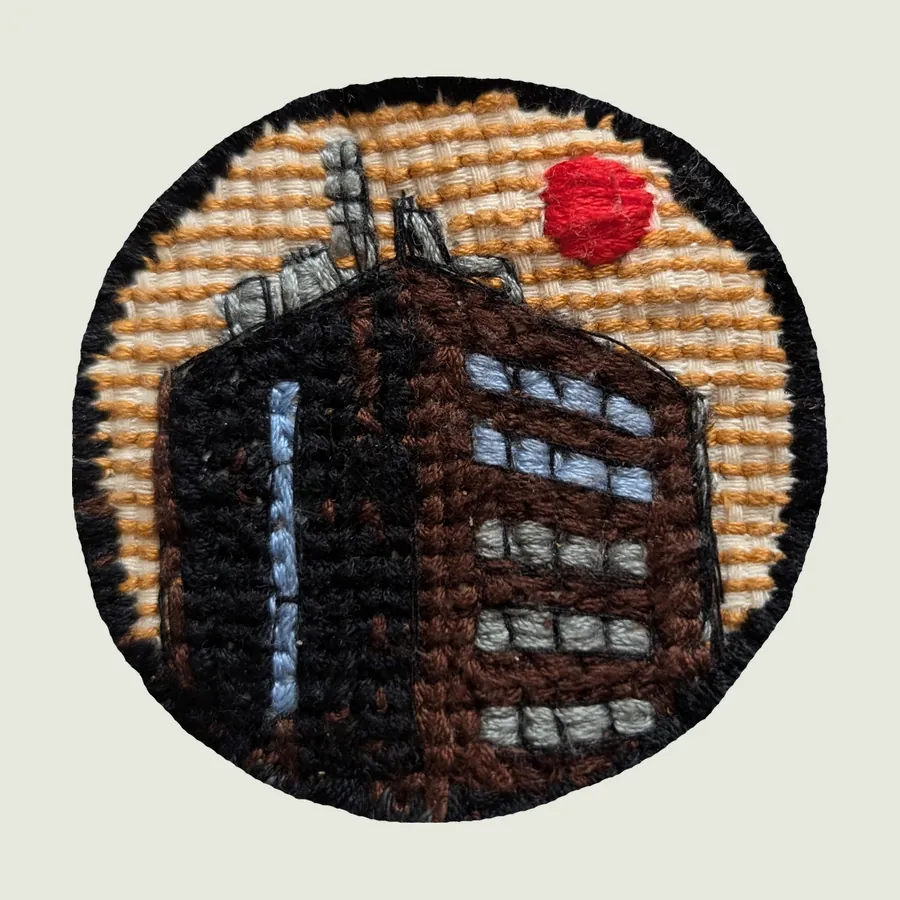 Round embroidered patch edged in black with white flecks: a brown and black tower block with pale windows against a pale yellow sky and a small red sun.