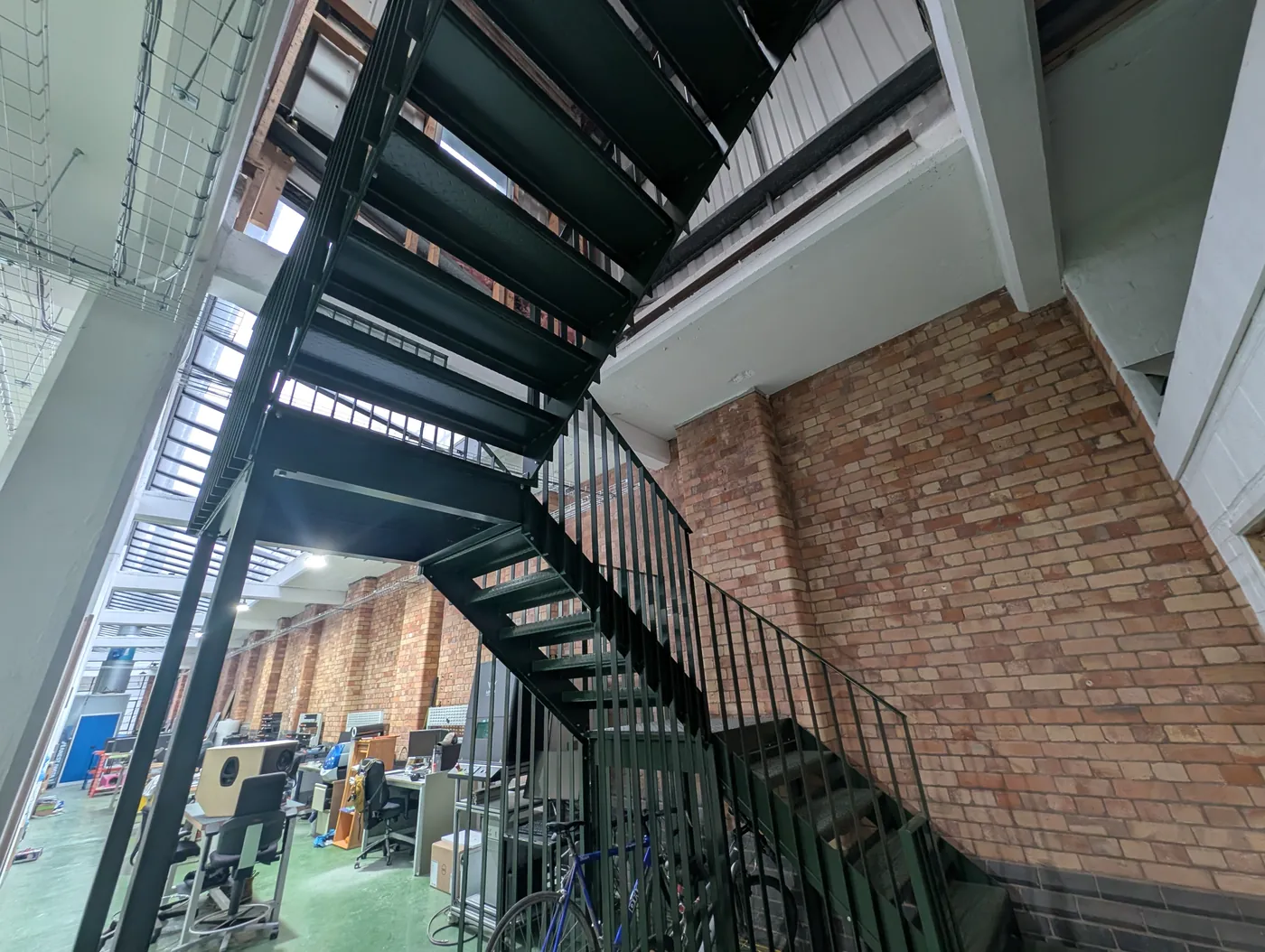 The same dark green staircase seen from below, its open treads and railings climbing along the brick wall, with workbenches on the floor beneath.