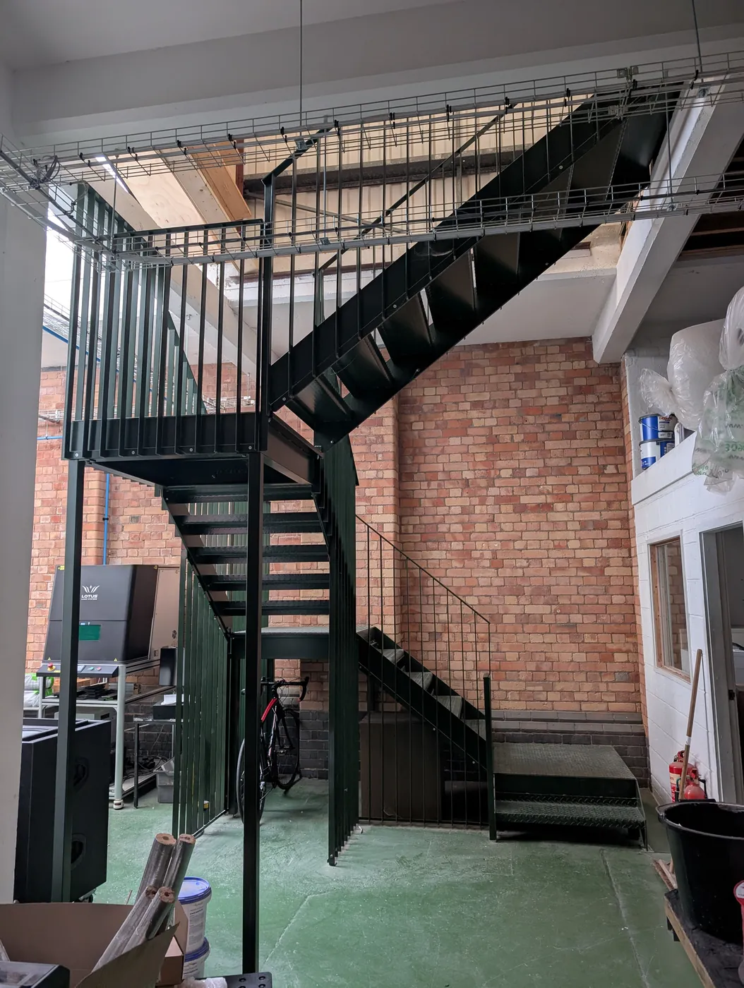 A dark green steel staircase in a factory: two flights of open treads with vertical-bar railings turning at a landing below a mesh-sided walkway, against a red brick wall.