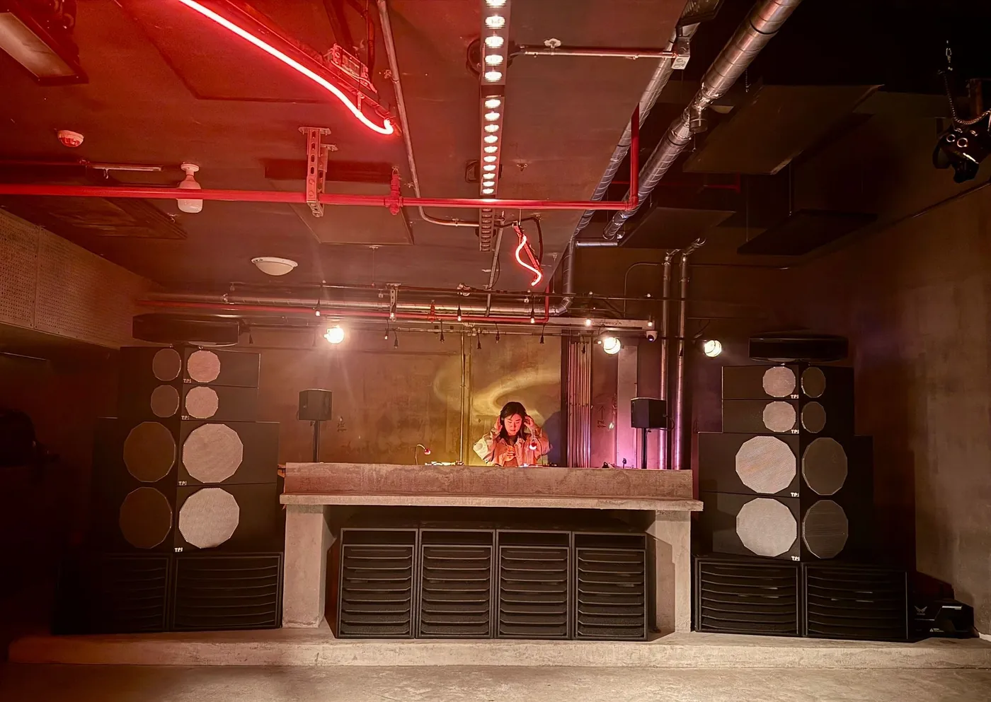 Two stacks of Type. TOTEM 2 in black, with grey octagonal grilles, either side of a DJ booth in a concrete club, red neon along the ceiling pipes.