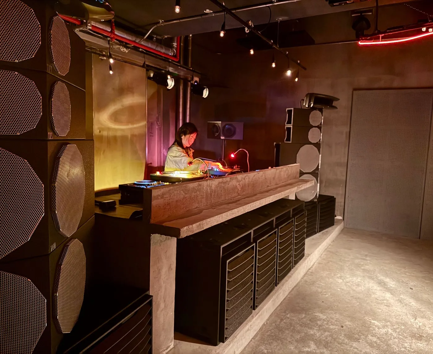 A black TOTEM 2 stack beside the DJ booth at Reactor: V-fronted modules with grey grilles above slatted bass cabinets, a DJ at the decks behind.