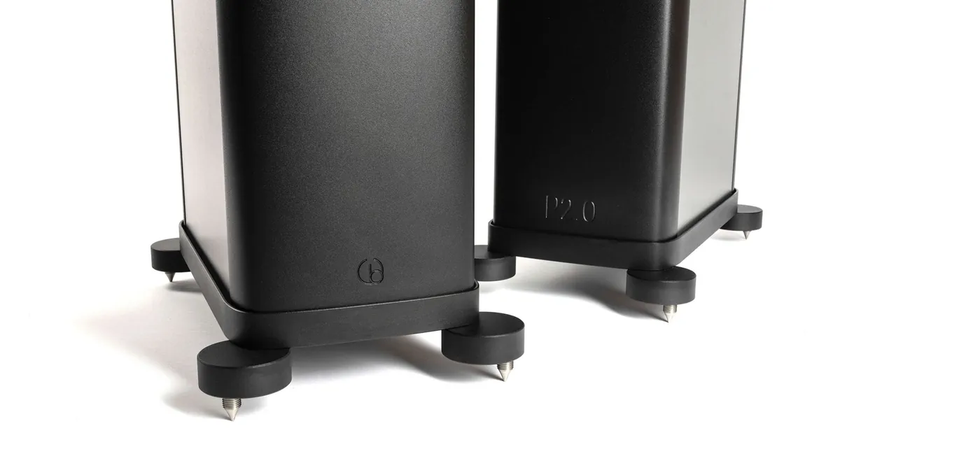 The bases of two black Precision P2.0 loudspeakers on white: machined bottom caps with round outrigger feet on steel spikes.